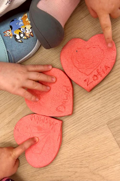 Na obrazku widać trzy różowe serca wykonane z materiału przypominającego gąbkę. Każde serce ma wypisane imię: "Daniel", "Mila" i "Zosia". Dziecięce rączki wskazują na serca, co sugeruje, że są one związane z jakąś zabawą lub aktywnością. W tle widać podłogę, która wygląda na drewnianą. Dziecięce buty są widoczne, co dodaje do całości przyjemnego, dziecięcego klimatu.